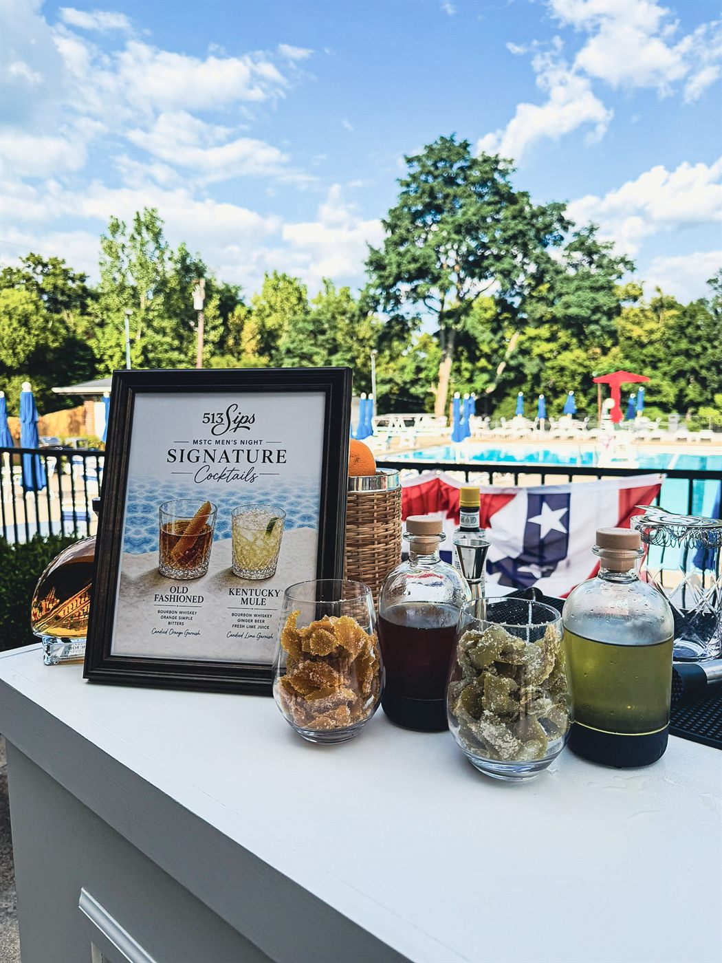 Two glasses of house-candied orange and lime peel beside bottles of house-made syrup on the 513Sips bar at Montgomery Swim and Tennis Club