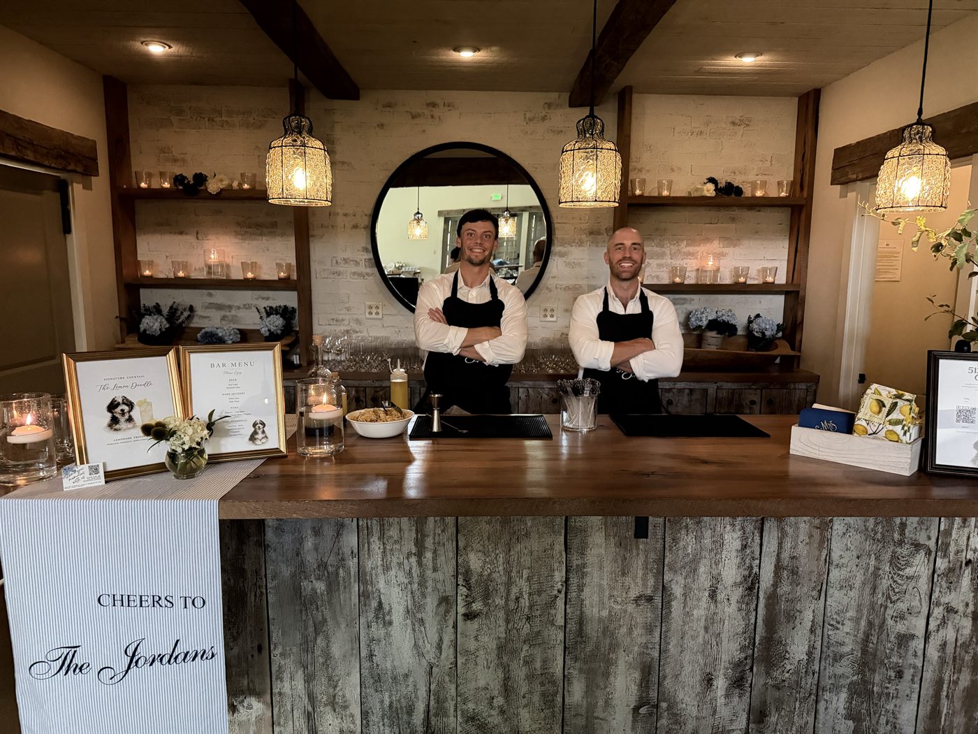 The full bar at Hope's Way dressed by 513Sips: framed signature cocktail and bar menu cards, candles, florals, garnish and printed napkins, with two bartenders behind it